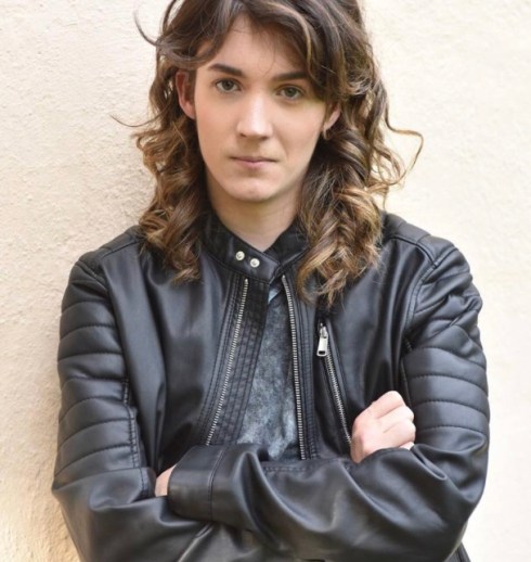 Mackenzie, with brown, curly hair with bangs, piercing brown eyes, and tan skin, stands against a stucco wall. Her arms are folded. She wears a black heather shirt with a leather jacket.
