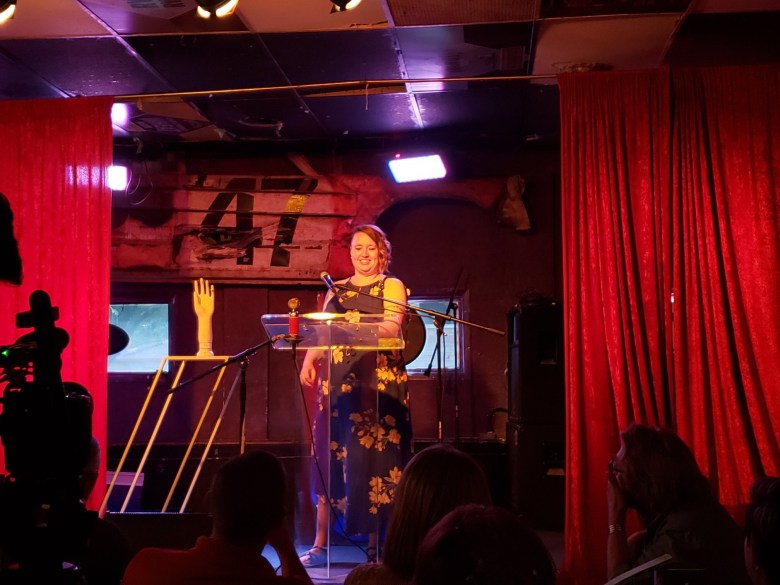 White woman with blonde hair, curls on right side, wearing a blue sleeveless dress with yellow flowers, stands on a stage behind clear podium, with microphone, on her right is a mannequin hand. There are red stage curtains to the left and right of the woman. The woman has a smiling expression on her face. In the audience, several people are looking toward the stage to see her.