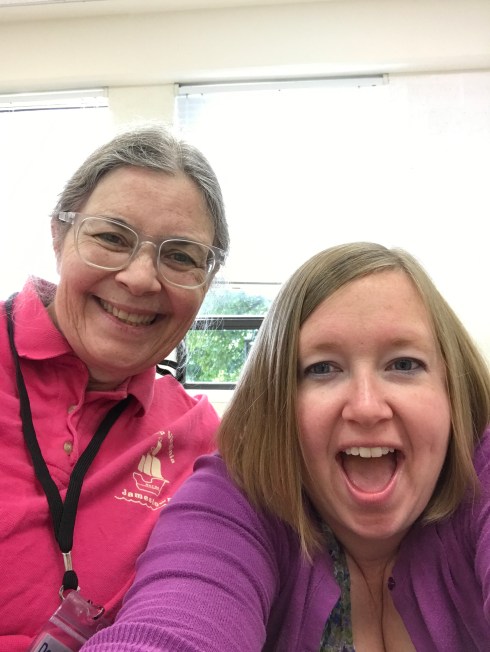 Two white women, standing in front of windows in a classroom, smile at the camera. The woman on the left has gray hair, parted down the middle and swept back into a ponytail. She's wearing clear-framed glasses, a pink "Cue Camp Virginia" polo shirt, and a black name tag lanyard. The woman on the right has chin-length blond hair, parted on the photo right side. Her mouth is open in a smile. She is wearing a flowery top and a purple cardigan sweater over it. Her arms are outstretched to take the selfie.
