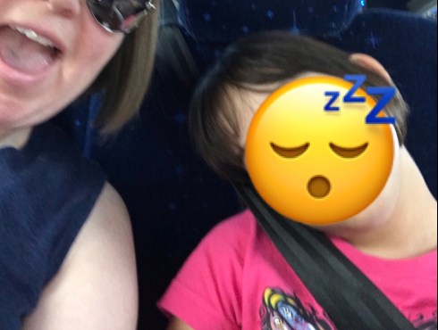 In the left side of the screen, a white woman is partially visible. she has blonde, chin-length hair, and is wearing mirrored silver sunglasses. Her mouth is open in a smile. She is wearing a blue, sleeveless t-shirt. Next to her is a little girl with brown, chin-length hair and brown skin. She is wearing a pink t-shirt with a colorful design on it. Her head rests on her shoulder as she sleeps. Her face is covered with a Zzz sleeping emoji to protect her identity. She is strapped into her seat with a black seat belt that goes across her body from her shoulder to her waist. They are in a travel bus.