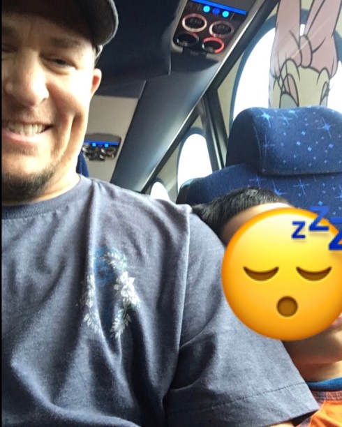 On the left side of the photo, three-quarters of the front of a man is visible. He is white, with a brown goatee. He is wearing a baseball cap and a blue t-shirt with a logo on the left (photo right) front of the chest. To the right, a little boy leans on him, sleeping. He has black hair and brown skin, and is wearing an orange sleeveless t-shirt with blue trim. His face is covered with a Zzz sleeping emoji to protect his identity. They are sitting in a travel bus.