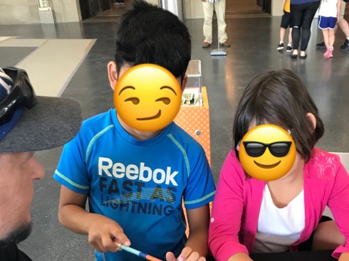 In the lower left corner is a man's face. He is white, with a brown goatee. He is wearing a baseball cap with a grey bill. Black sunglasses are perched atop the hat bill. Next to him, a little boy stands. He has black hair and brown skin. He is wearing a short-sleeved, blue, Dri Fit t-shirt that says, "Reebok" in white text, then, "Fast as lightning" below, in black text. The shirt has white stitching at the seams. His face is covered with a slanted smile emoji, to protect his identity. Next to him, a girl leans on the table. She has brown hair and brown skin. She is wearing a white t-shirt with a pink cardigan over it. Her face is covered with a smiling sunglasses emoji, to protect her identity. In the back ground, there are some people standing in the top right of the photo. They are only visible from the waist down.