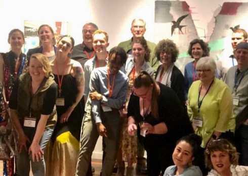 Approximately 20 people stand in three rows, posing and smiling for the picture. In the lower right corner of the photo, two women sit on the ground and look at the camera.